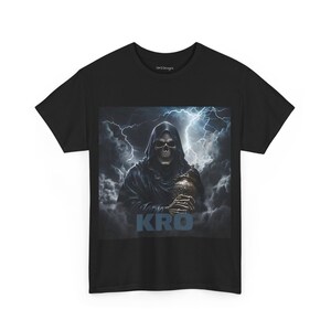May include: Black t-shirt with a graphic print of a hooded skeleton holding a skull in a stormy sky. The text "KRO" is printed below the skeleton.