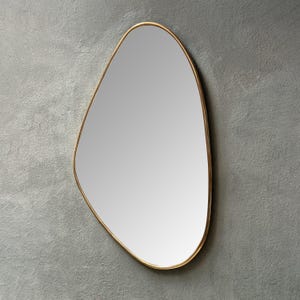 Puede incluir: Un espejo de forma irregular con un marco de metal dorado. La superficie reflectante del espejo es lisa y clara, sobre un fondo gris. El marco tiene un perfil delgado, agregando un toque moderno al diseño.