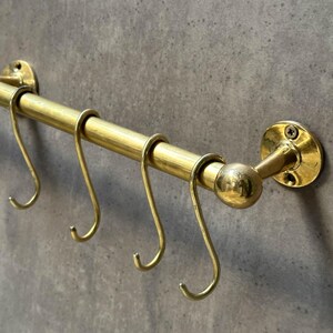 May include: Brass pot rack with five hooks. The rack is mounted to a wall with a round base and a decorative ball finial.