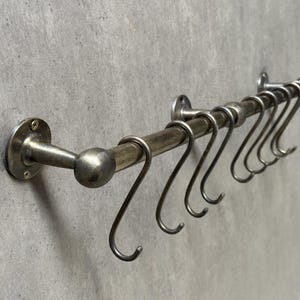 Puede incluir: Un estante para utensilios de cocina de latón con una fila de ganchos. El estante está montado en una pared y tiene una tapa redonda decorativa.