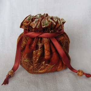 May include: A small drawstring bag with a red ribbon and gold accents. The bag is made of a fabric with a gold and orange leaf pattern.