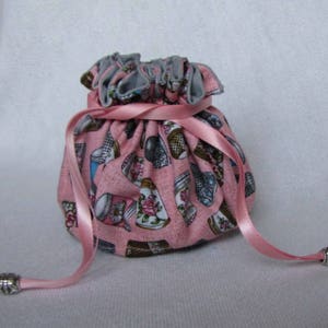 May include: A pink drawstring bag with a gray lining and a pink ribbon. The bag is decorated with a pattern of teacups and saucers.
