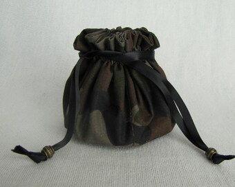 Puede incluir: Una pequeña bolsa redonda con cordón y un patrón de camuflaje en tonos marrón, verde y negro. La bolsa tiene un cierre de cordón de cinta negra con cuentas decorativas de color bronce en los extremos.