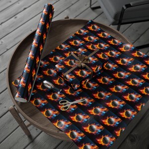 Op de afbeelding: Een rol cadeaupapier, een ingepakt cadeau en een vel cadeaupapier met een herhaald patroon van een vlammende basketbal op een donkerblauwe achtergrond. Een schaar en plakband zijn ook aanwezig.