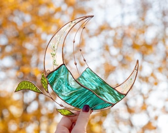 May include: A teal blue and copper stained glass crescent moon with a delicate floral design. The moon is suspended from a copper wire and has a leafy stem extending from the bottom.