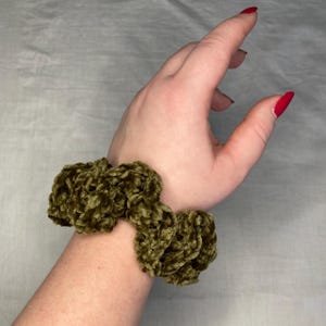 Può includere: Un elastico per capelli in velluto verde con una texture soffice. L'elastico è indossato al polso.