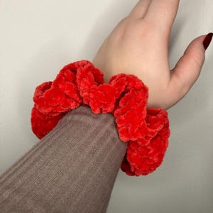 Può includere: Un elastico per capelli rosso e soffice fatto di filato morbido e soffice. L'elastico è legato intorno a un polso.