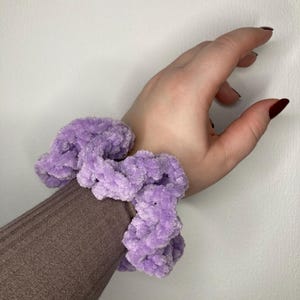 Può includere: Un elastico per capelli viola e peloso fatto di filato. Lo scrunchie è indossato al polso.
