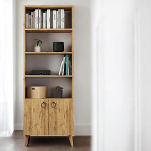Op de afbeelding: Een hoge, lichtgekleurde houten boekenkast met meerdere planken en een onderkast. De planken tonen boeken, decoratieve items en kleine planten. De kastdeuren hebben ringtrekkers. De boekenkast staat tegen een witte muur.