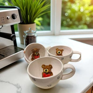 Puede incluir: Tres tazas de café de cerámica blanca con figuras de osos que sostienen corazones rojos en su interior. Cada taza tiene un asa y diseños de huellas de patas. Una máquina de espresso plateada está en el fondo.