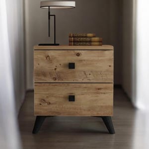 May include: A two-drawer wooden bedside table with a natural finish. The bedside table has black square knobs and black legs. A black desk lamp with a white lampshade and books sit on top of the table.