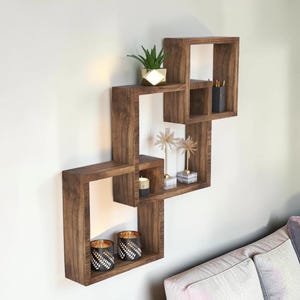 May include: A wall-mounted shelving unit composed of interconnected dark brown wooden square frames. The shelves feature a natural wood grain finish and hold various decorative items including a small green plant, candles, and metallic ornaments.