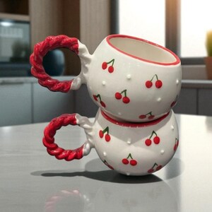 Puede incluir: Dos tazas de cerámica blancas apiladas, adornadas con cerezas rojas y pequeños puntos blancos. Las asas tienen un diseño rojo trenzado. Las tazas están sobre una superficie gris claro, lo que sugiere una cocina.