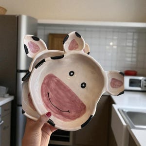 May include: A stack of two ceramic cow-shaped bowls. The bowls are white with pink and black accents, featuring a pink snout, black eyes, and a smiling mouth. The ears are also pink with black spots. The bowls are held in a kitchen setting.