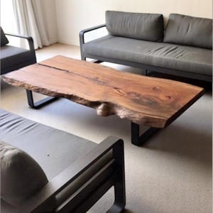 May include: A large, rustic wooden coffee table with a live edge top and black metal legs. The table is in a living room setting with a gray sofa in the background.