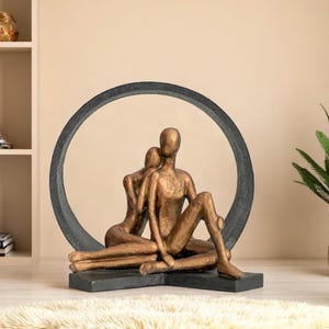 Puede incluir: Escultura de bronce de dos figuras abrazándose, sentadas frente a un marco circular gris oscuro. La escultura está sobre una base gris oscuro, y las figuras están estilizadas con formas lisas y sin rasgos. La escena evoca intimidad y conexión.