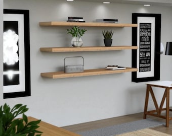 May include: Three wooden floating shelves mounted on a light gray wall. The shelves hold books, plants in glass and black pots, and decorative items. Two framed art pieces are on either side of the shelves. A small table and lamp are in the background.