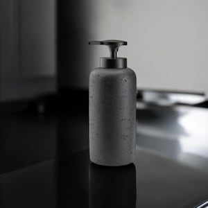 May include: A dark gray soap dispenser with a matte finish. The cylindrical bottle has a textured surface and a black pump. The dispenser is designed for liquid soap and is suitable for a bathroom or kitchen.