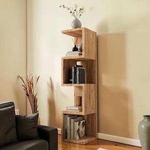 Könnte beinhalten: Ein hohes Bücherregal aus hellem Holz mit einem Zickzack-Design in S-Form und vier offenen Ablagefächern. Die Regale sind mit verschiedenen Büchern, einer kleinen schwarzen Vase und einer grauen Vase mit getrockneten Blumen gefüllt.