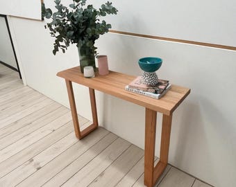 Könnte beinhalten: Eine hellbraune Holzkonsolentisch mit rechteckiger Platte und abgewinkelten Beinen. Der Tisch ist mit einer Vase mit Grün, Kerzen, Büchern und einer türkisfarbenen Schale dekoriert. Der Tisch ist 122 cm lang.