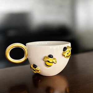 May include: A white ceramic teacup with a gold handle and three bee embellishments. The bees are yellow and black with gold wings. The teacup is on a dark brown surface.