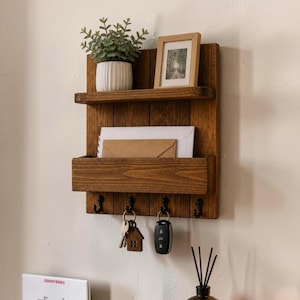 May include: A wooden wall-mounted organizer with a shelf, a mail slot, and key hooks. It holds a small potted plant, a framed picture, and envelopes. Keys and a car key are hanging from the hooks. The wood has a warm, brown finish.