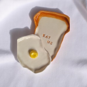 May include: Ceramic breakfast-themed dish set. The set includes a toast-shaped plate with the words "EAT ME" and a separate egg-shaped plate with a yellow yolk. The plates are off-white with brown and yellow accents.