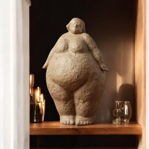 May include: A beige ceramic sculpture of a standing person with a rounded body shape. The sculpture is displayed on a wooden shelf.