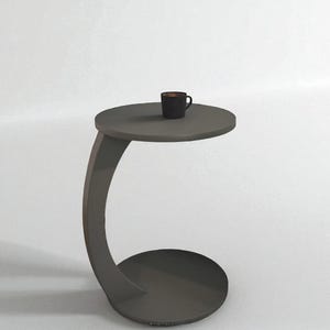Pode incluir: Uma mesa lateral cinza moderna com uma perna curva e um tampo e base redondos. Uma pequena xícara de café preta está sobre a superfície da mesa. A mesa tem aproximadamente 51 cm de altura.