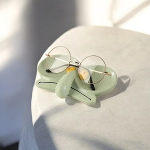 May include: A green ceramic glasses holder shaped like Squidward from Spongebob Squarepants. The holder has a yellow nose and black eyes. A pair of round glasses is resting on the holder.