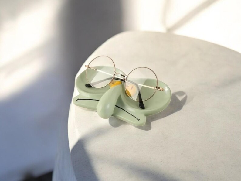 May include: A green ceramic glasses holder shaped like Squidward from Spongebob Squarepants. The holder has a yellow nose and black eyes. A pair of round glasses is resting on the holder.