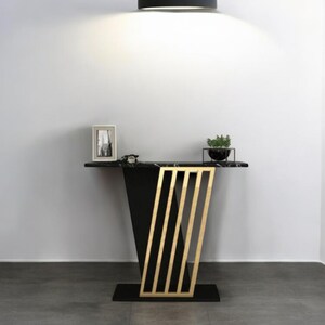 Könnte beinhalten: Ein moderner schwarzer Konsolentisch mit Marmorplatte und goldenen Akzenten. Der Tisch hat ein geometrisches Design mit vertikalen goldenen Stäben auf der Vorderseite. Ein kleines gerahmtes Bild und eine Topfpflanze stehen darauf.