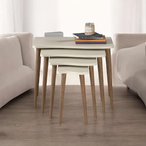 May include: A set of three nesting tables with a light beige finish and wooden legs. The tables are arranged in descending order of size, with the largest table at the top. Books and a candle sit on the largest table. The tables are surrounded by two beige sofas.
