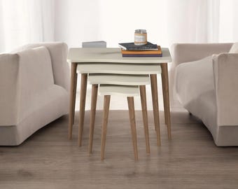 Puede incluir: Un juego de tres mesas nido con acabado beige claro y patas de madera. Las mesas están dispuestas en orden descendente de tamaño, con la mesa más grande en la parte superior. Libros y una vela están sobre la mesa más grande. Las mesas están rodeadas por dos sofás beige.