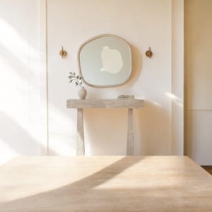 May include: A neutral-toned interior scene featuring a light wood console table with a decorative mirror. The mirror has an organic shape and a pale green interior design. A small vase with greenery and two brass wall sconces complete the decor.