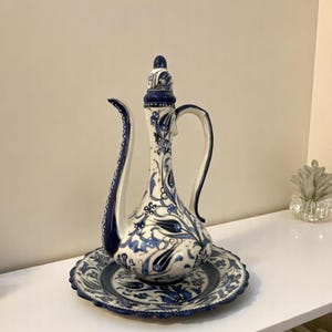 Könnte beinhalten: Eine hohe, verzierte Keramikkaraffe mit passendem Teller. Die Karaffe und der Teller sind weiß mit einem blauen Blumenmuster. Die Karaffe hat einen langen Ausguss und einen gebogenen Griff. Dekorative Tischware.