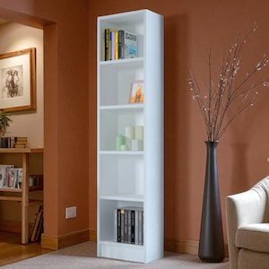 May include: A tall, white bookshelf with multiple shelves, filled with books, candles, and decorative items. The bookshelf is positioned against a warm-toned wall, with a tall, dark vase holding branches nearby. A framed picture and a small table are also visible.