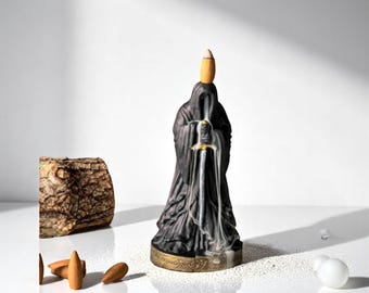 May include: Black ceramic incense burner shaped like a figure in a long robe holding a sword. The incense cone is placed on the figure's head and smoke flows down the sword.