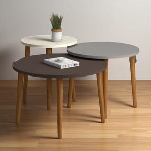 Puede incluir: Tres mesas auxiliares redondas en varios colores: marrón oscuro, gris claro y blanco roto. Cada mesa tiene patas de madera y un diseño minimalista. Una pequeña planta en maceta está sobre la mesa blanca.