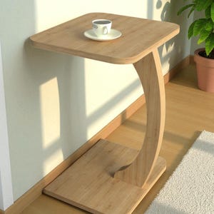 Pu&ograve; includere: Un tavolino in legno chiaro con piano quadrato e gamba curva. Una tazza e un piattino bianchi sono appoggiati sul tavolo. Il tavolo &egrave; vicino a un muro e a una finestra, con una pianta in vaso sullo sfondo.