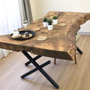 May include: A large wooden table with a live edge top and black metal X-shaped legs. The table is decorated with small potted plants and coasters.