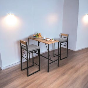May include: A small, modern dining set with a light wood table and two matching bar stools. The table is set with a plate of food, a cup, and a bowl of fruit. The stools have gray upholstered seats and black metal frames.