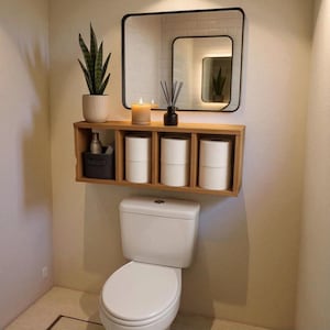 Könnte beinhalten: Ein modernes Badezimmer mit einer weißen Toilette, einem Holzregal mit Toilettenpapierrollen und einem schwarz gerahmten Spiegel. Eine Topfpflanze und eine Kerze sorgen für dekorative Akzente. Der Raum hat eine neutrale Farbpalette.