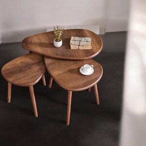 Op de afbeelding: Set van drie houten nesttafels met een natuurlijke afwerking. De tafels hebben een organische vorm en worden ondersteund door drie taps toelopende poten. Een klein wit kopje en schotel staan op de kleinste tafel.