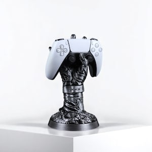 Può includere: Un controller di gioco bianco e nero poggia su un supporto nero a forma di mano. Il supporto ha un design strutturato e dettagliato e una base circolare. Il controller è bianco con pulsanti neri.