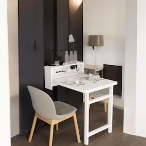 May include: A white, foldable dining table with a matching chair and a built-in cabinet. The table is set with glassware and cutlery. The chair has a gray seat and wooden legs. The cabinet is dark gray.