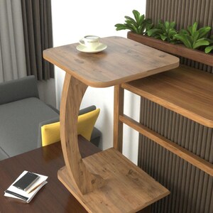 Pu&ograve; includere: Un tavolino in legno con una gamba curva e due livelli. Una tazza di caff&egrave; &egrave; appoggiata sul livello superiore. Il tavolo &egrave; in legno marrone chiaro e ha un design moderno. Il tavolo &egrave; alto circa 61 cm.