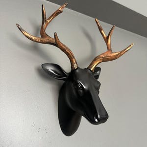 Puede incluir: Una cabeza de ciervo negra para montar en la pared con cornamenta de color bronce. La cabeza de ciervo es brillante y tiene un acabado liso. La cornamenta tiene un aspecto texturizado y es de un color metálico cálido. Esta pieza decorativa está diseñada para la pared.