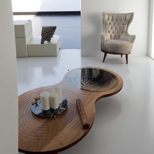 Puede incluir: Una escena de salón moderno con una mesa de centro de madera única con tapa de cristal y un arreglo decorativo de velas. También se ve un sillón con mechones y una escultura estampada.