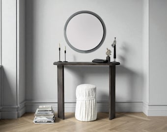 Puede incluir: Escena interior moderna con una consola de madera oscura y un espejo redondo. La mesa exhibe objetos decorativos, incluyendo candelabros y una escultura. Debajo hay un taburete blanco texturizado.